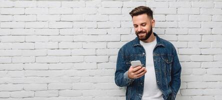 The young man happily checking his smartphone against a white brick wall. photo