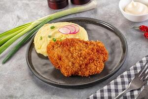 A plate with a fried chicken cutlet, mashed potatoes, and a side of pickles photo