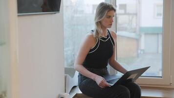 Young professional working remotely, sitting by window in bright modern apartment, using laptop with focused concentration on digital task video