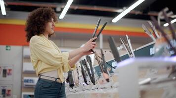 Young woman carefully comparing hair straightener features while browsing electronic store display, evaluating product details with focused concentration in slow motion video