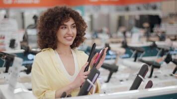 customer exploring hair styling technology. Comparing multiple hair straightener models while smiling confidently in electronics store retail environment during slow motion shopping experience video