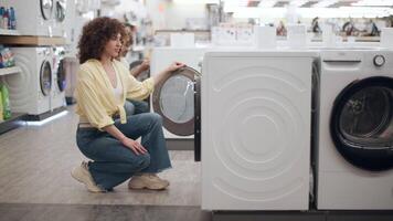 Young female shopper examining newly displayed washing machine, browsing domestic appliance selection with satisfied smile in modern electronics store environment video