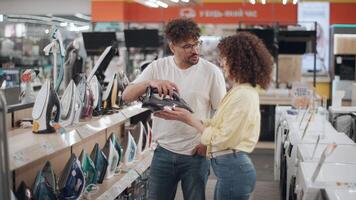 Young couple browsing household appliances in sleek electronics store, carefully comparing iron features and evaluating options with focused shopping energy in smooth slow motion video