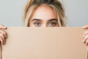 A young woman with blue eyes holds a blank cardboard sign in front of her face against a light background photo