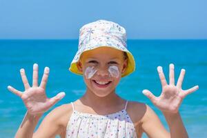 Happy girl at the beach, wearing a colorful sun hat, with sunscreen applied on her face and hands raised, enjoying summer fun and promoting sun safety on blue sea photo