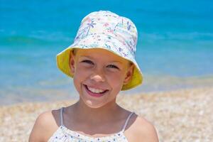 Smiling girl in a vibrant sun hat enjoys a sunny day at the beach, with soft sand and clear turquoise water in the background, capturing a moment of happiness photo
