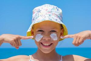 Caucasian smiling child of 8 9 years in a sun hat showing on sunscreen on cheeks over sea looking at camera photo