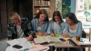 caucasiano quatro adolescentes escrevendo dever de casa preparando colegas de classe aprender dentro biblioteca escola alunos alunos com livro preparar exames dentro Academia Faculdade estude juntos Aprendendo Educação falando trabalho em equipe video