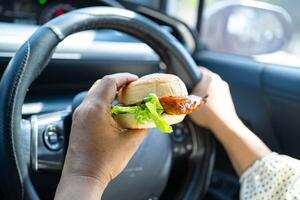 Asian woman driver hold and eat hamburger in car, dangerous and risk an accident. photo