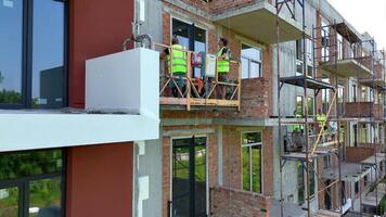 Construction workers on suspended platform installing facade panels, New apartment block under active exterior finishing and thermal insulation phase video