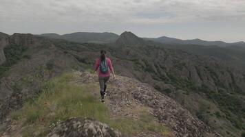 Aerial view Female hiker walking alone along rocky mountain trail, surrounded by dramatic ridges and valleys under overcast sky, symbolizing solitude, exploration, and physical resilience in nature video