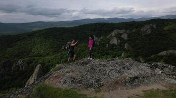 Aerial view active Couple hiking on rocky mountain reaching top enjoy views healthy lifestyle and outdoors climb to summit with lush green forest and cloudy sky in background video