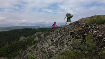 aerial view Man helps woman climb rocky trail on mountain slope with lush forest and cloudy sky in background, teamwork and adventure concept video