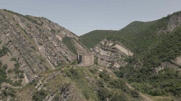 Ancient stone fortress ruin on mountain ridge surrounded by forested valleys and distant peaks in summer daylight, aerial view video