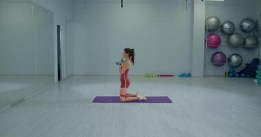 Girl athlete performs kneeling squat exercise with dumbbells in a well lit gym studio during a fitness session video
