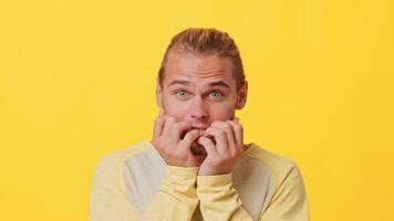 A young guy with mixed ancestry displays a nervous expression by biting his nails while standing in front of a bright yellow backdrop. The mood captures a relatable moment of anxiety. video