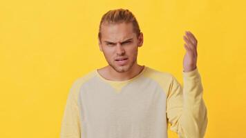 A young man with a mixed heritage shows frustration while gesturing with his hands. The vibrant yellow backdrop adds intensity to his puzzled expression in a casual setting. video