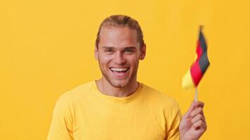 A young man with a bright smile is showing German flag in his hand. He is wearing a yellow shirt and stands in front of a vivid yellow background. The mood is cheerful and lively. video