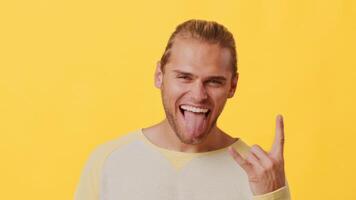A young man with a mix of African and Caucasian features smiles widely, sticking out his tongue while making a rock hand gesture. The vibrant yellow background adds energy to the moment. video
