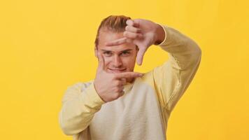 A young man playfully poses in front of a vibrant yellow backdrop, using his hands to create a frame. His confident expression reflects a sense of creativity and energy typical of Generation Z. video