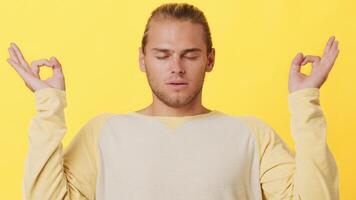 A young man with a calm expression practices meditation while seated with hands in a peaceful position. He is dressed in a light sweater, exuding serenity in a vibrant yellow setting. video