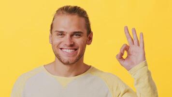 A cheerful young man with a stylish haircut smiles warmly while making an OK gesture. He stands in front of a bright yellow background, giving off positive energy and confidence. video