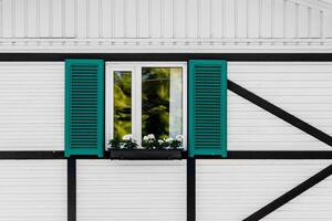 A window with a white frame, green shutters, and a box with white flowers on the facade of a house with white siding and black beams. photo