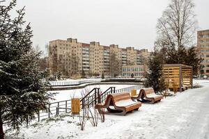 A snow-covered city park with benches and a fir tree against the backdrop of a typical Soviet residential building on a cloudy winter day. photo