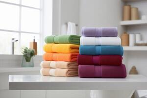 Two stacks of colorful towels on a white table in the bathroom, a window and shelves with items in the background. photo