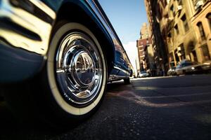 A retro car wheel with a shiny hubcap and a white tire on asphalt, buildings in the background. photo