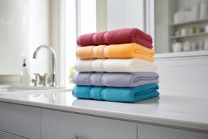 A stack of five folded towels of different colors on a white countertop in a bathroom, a faucet is visible. photo
