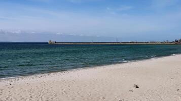 Scenic view of a quiet sandy beach and calm sea waves under a clear blue sky. A breakwater structure with buildings and a watchtower stretches along the horizon in the distance. photo