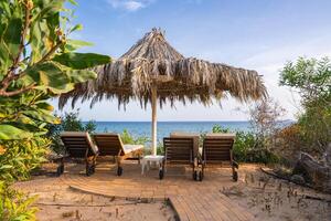 Four wooden lounge chairs sit under a thatched umbrella on a paved area, surrounded by greenery, with a clear view of the sea and horizon in Cyprus. photo