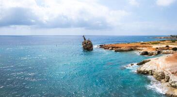The Edro III Shipwreck rests tilted in turquoise waters near Cyprus, surrounded by rocky cliffs, a reddish brown shoreline, and a partly cloudy sky. photo
