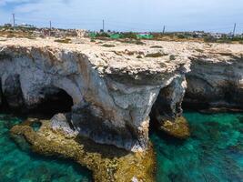 Rocky cliffs with natural sea caves meet turquoise waters in Ayia Napa, Cyprus. A sculpture park and scattered buildings are visible in the background. photo