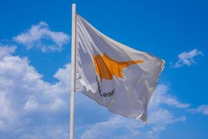 The national flag of Cyprus waves against a clear blue sky with scattered white clouds, featuring a golden island silhouette and green olive branches. photo
