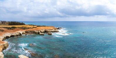 Rugged cliffs meet clear turquoise waters along the Mediterranean coast in Cyprus. Sparse vegetation and distant buildings enhance the seascape. photo