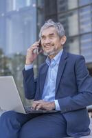 senior professional business man typing laptop computer and using smartphone talking to client while sitting outside building photo
