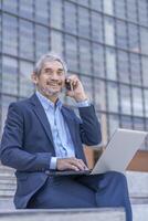 senior professional business man typing laptop computer and using smartphone talking to client,sitting outside building photo