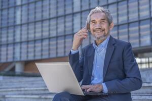 busy senior professional business man typing laptop computer and using smartphone talking to client while sitting outside building photo