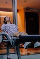 A woman is using a leg extension machine in a gym. Her legs are positioned on the machine. The gym has large windows and a door can be seen in the background. photo