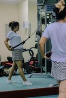 A woman uses a cable machine in a gym, doing an exercise routine. She is seen from the side. Her reflection can be seen in the mirror, showing her front and back views. photo