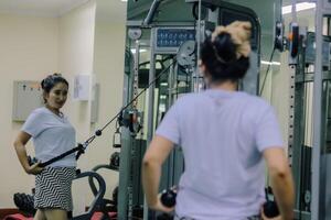 A woman uses a cable exercise machine at a gym. She is facing the machine, but there is also a mirror reflection showing her side profile. photo
