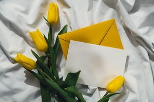 Mock up of white empty envelope greeting card with yellow fresh tulips on white bed with trendy deep shadows. Spring flowers in yellow colors. Romantic postcard wallpaper design Copy space photo