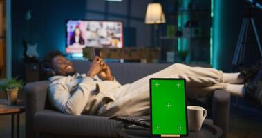 African american young adult laying on sofa next to mockup copy space, relaxing and browsing web on smartphone. Happy relaxed person on couch feeling happy with remote job. Camera B. photo