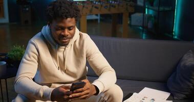 African American man on sofa texting mates in living room, entertained by social media and messages. Depiction of remote work breaks, digital communication and relaxation at home. Camera B. photo