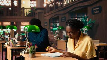 African american pupil highlighting key words from class notes, creating summary to study easily and memorize the lesson theory. Student using learning technique in library. Camera A. photo