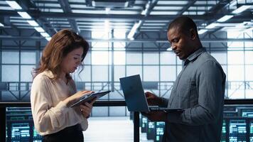 Developers using laptop and tablet, executing security patches and securing firewall. Server farm workers using devices to perform vulnerability scans, ensuring data protection, camera B photo