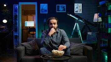 Puzzled man eating popcorn while watching confusing movie on streaming service, enjoying time off from work. Person consuming snacks, looking at film broadcasted on TV, camera B photo