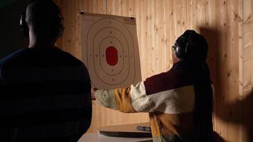 Woman in firing range training with pistol, looking at results on paper target. African american person doing shooting sport, sharpening skills and checking points scored on bullseye, camera A photo
